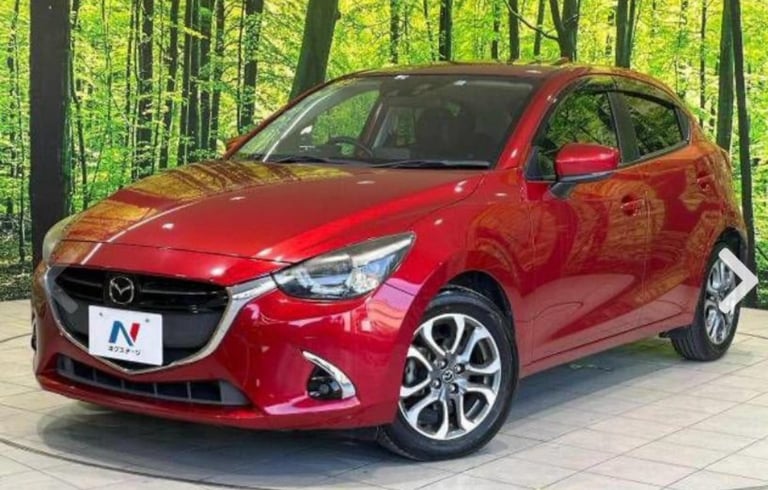 Red Mazda compact hatchback parked on a paved surface with green forest in the background